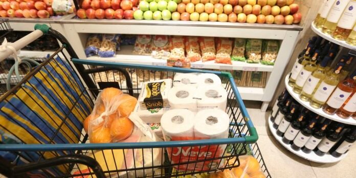 Grocery cart in a supermarket filled with toilet paper, onions, and packaged groceries, with produce and wine shelves in the background