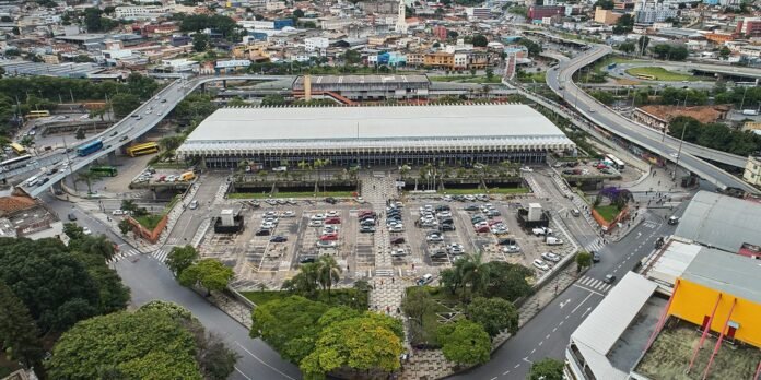 Aerial view of a large white-roofed building surrounded by a landscaped plaza and a busy parking lot, with roads and city buildings around it.