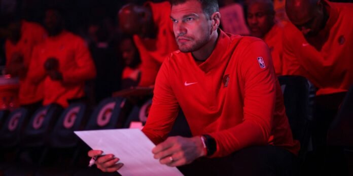 Basketball player in a red warm-up shirt sits at a table, holds a clipboard and marker, looking focused in a dim red-lit arena audience.