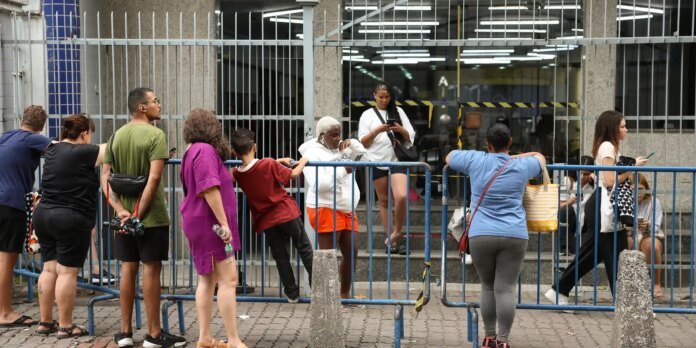 People line up behind blue metal barriers outside a building entrance, waiting or chatting.