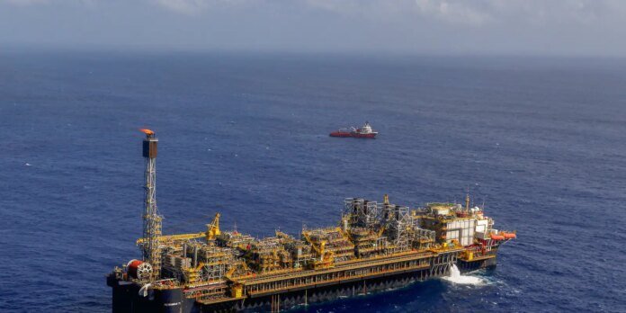 Offshore oil drilling platform with yellow scaffolding and machinery, in the open sea with a small support vessel in the distance.