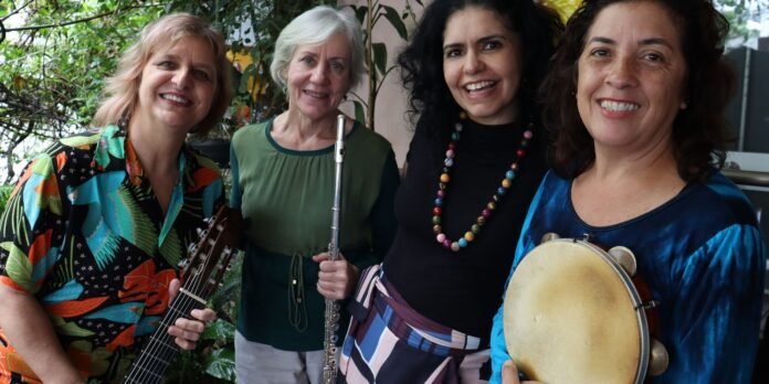 Four women musicians smiling outdoors: left holding a guitar, second with a flute, third wearing colorful bead necklace, right with a tambourine.