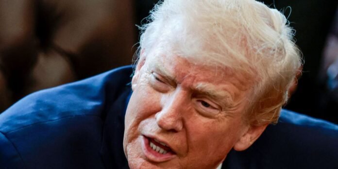 Close-up of a light-haired man grimacing, eyes squinting, teeth showing, in a dark blue suit.