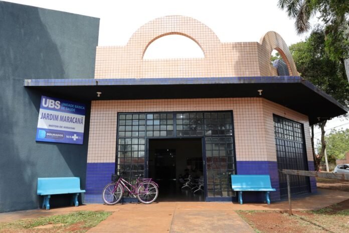 Health clinic entrance with pink tile exterior, blue benches, and a pink bicycle parked by the doorway; sign reads UBS Jardim Maracanã.