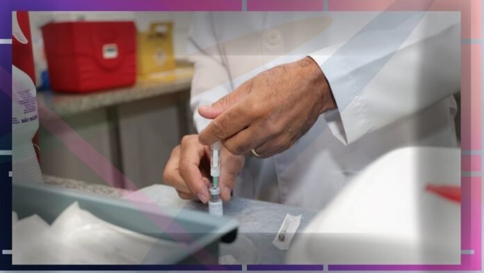 Clinician in a white coat drawing up medication from a vial with a syringe at a lab bench.