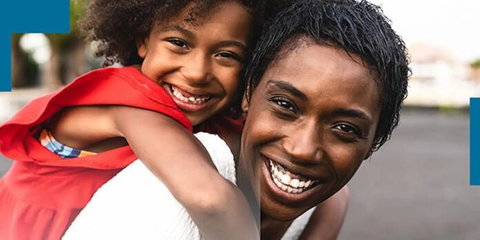 Joyful woman with a child on her back, both smiling for the camera outdoors.