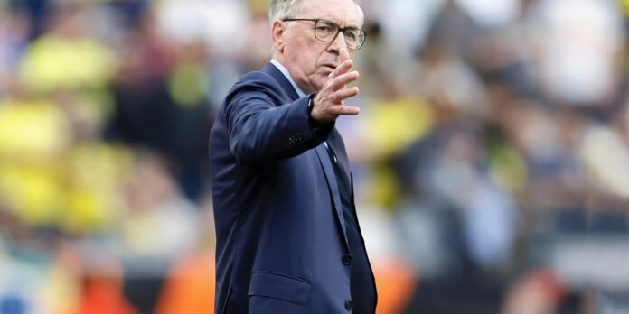 Man in a navy suit and glasses gestures toward camera on a sports field with blurred crowd in the background