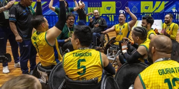 Wheelchair basketball team in yellow uniforms celebrates in a huddle on a wooden court, smiling and raising arms amid cheering teammates and coaches.