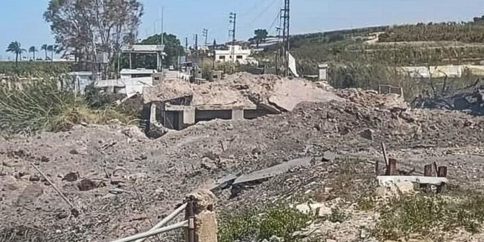 Rubble-strewn construction site with a partially collapsed concrete structure and exposed foundation, power lines and distant hills in the background.