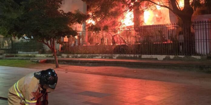 Firefighter kneels on a sidewalk as a building behind a fence burns with bright flames at night.