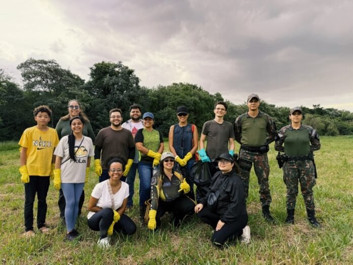 Diverse group of volunteers posing in a grassy field with trees in the background, all wearing gloves for an outdoor cleanup activity and smiling at the camera.