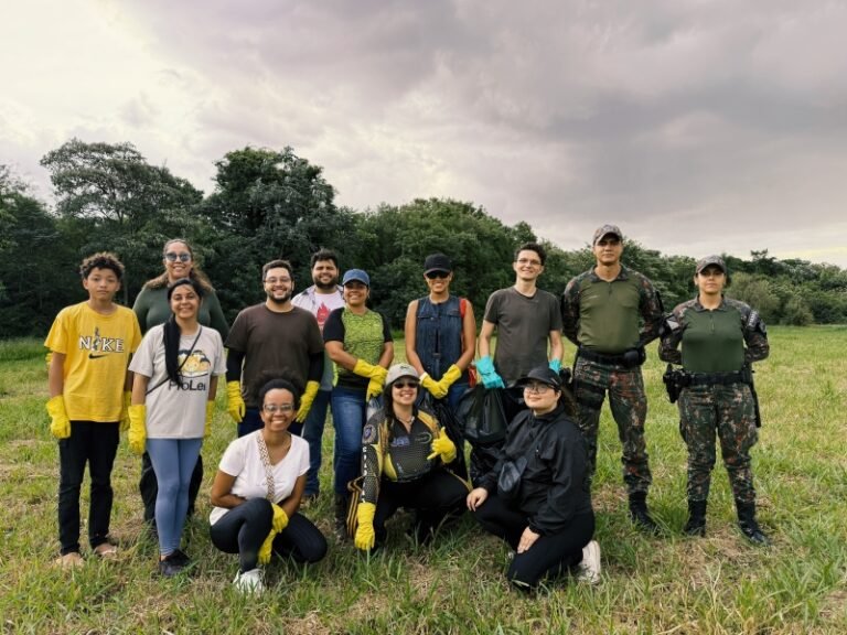 Diverse group of volunteers posing in a grassy field with trees in the background, all wearing gloves for an outdoor cleanup activity and smiling at the camera.
