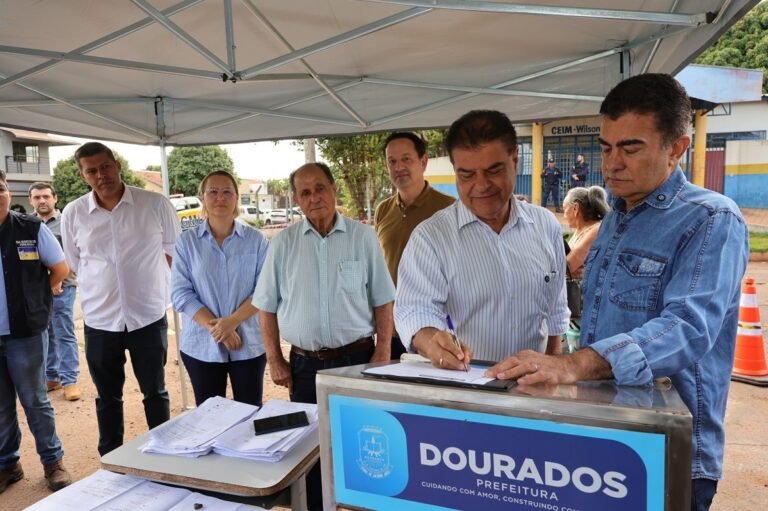 Officials sign documents at an outdoor municipal event under a shelter, Dourados Prefeitura banner in view.