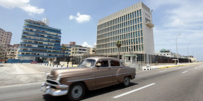 Brown vintage car drives along a sunlit city street with a blue curved residential building on the left and modern beige offices on the right.