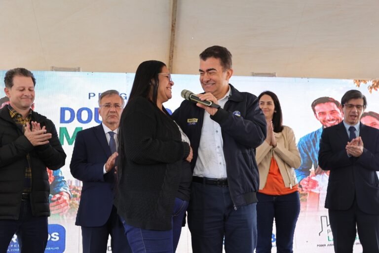 Man hands a microphone to a smiling woman on stage as colleagues clap behind them at a ceremony.