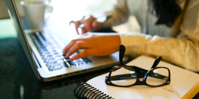 Person typing on a laptop at a desk, with glasses resting on a spiral notebook in the foreground.