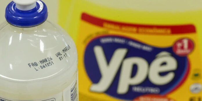 Close-up of a white plastic detergent bottle with a blue cap next to a yellow detergent box with a large blue logo in the background