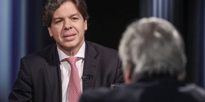 Man in a dark suit with a pink patterned tie speaks during an interview, microphone clipped to his lapel, facing an interviewer.