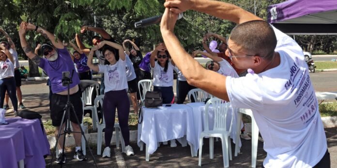 Group of people outside doing arm stretches during a fitness event, led by a man with a microphone on the right.