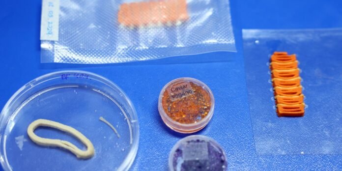 Lab tabletop with a small round container of orange vegan caviar labeled “Caviar Vegane” at center; a curved pale dough-like strip in a petri dish to the left; orange stacked pieces on the right; blue background with a foil packet overhead.