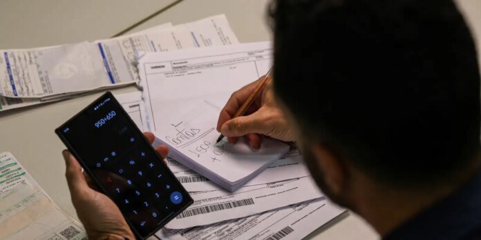 Person uses a smartphone calculator while writing totals on a notepad placed on top of scattered invoices and receipts.