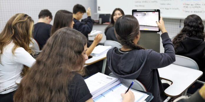 Group of students in a classroom; a girl holds up a tablet to record while others take notes in notebooks.