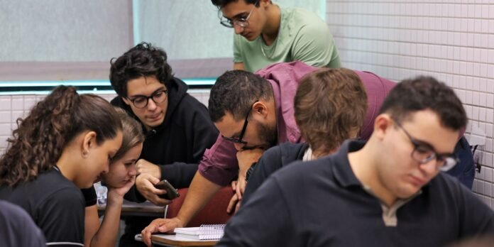 Group of students leaning over a notebook on a desk, collaboratively discussing in a classroom setting.