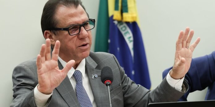 Man in a gray suit and glasses speaks into a microphone, hands raised, with a Brazilian flag in the background.