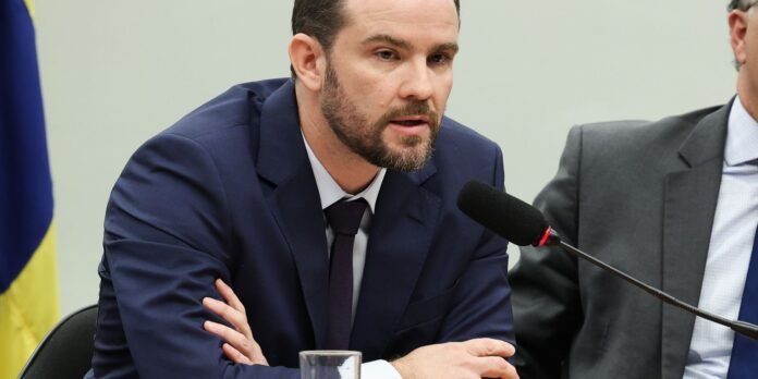 Man in a navy suit speaking into a microphone at a panel, with a second man in a gray suit nearby and a flag visible behind them