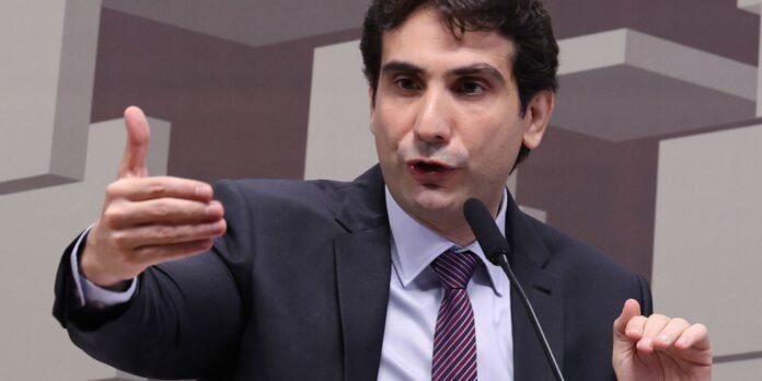 Man in a dark suit and striped tie speaking into a microphone, gesturing with one hand extended.