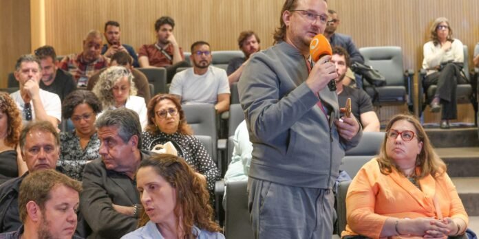 Man with glasses and shoulder-length hair speaks into an orange microphone while standing in a conference hall, audience seated and listening behind him.