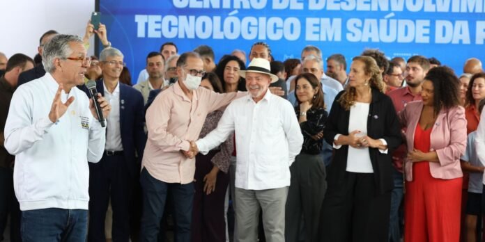 Speaker with microphone addresses a crowd at a tech-health event; grup of people listening and shaking hands in front of a blue banner.