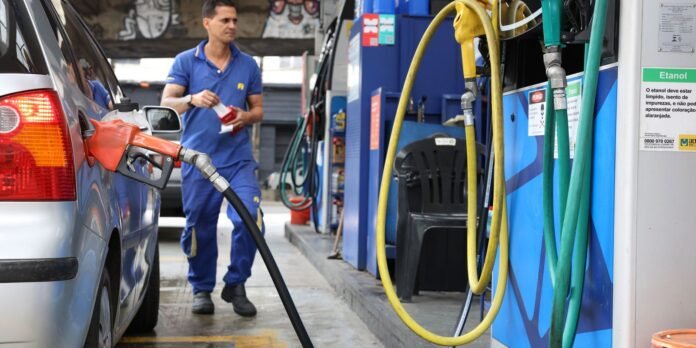 Gas station attendant in blue uniform refueling a silver car, with yellow and green hoses nearby.