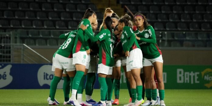 Women’s soccer players in green uniforms huddle together on the field during a match.