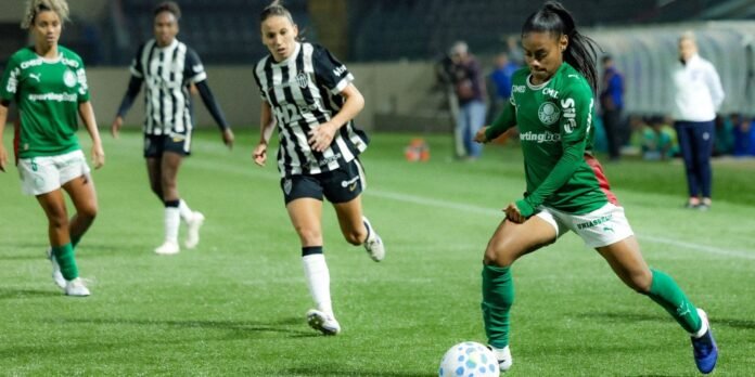 Female soccer player in a green Palmeiras kit controls the ball while an opponent in a black-and-white striped jersey pursues nearby on a well-lit field at night.