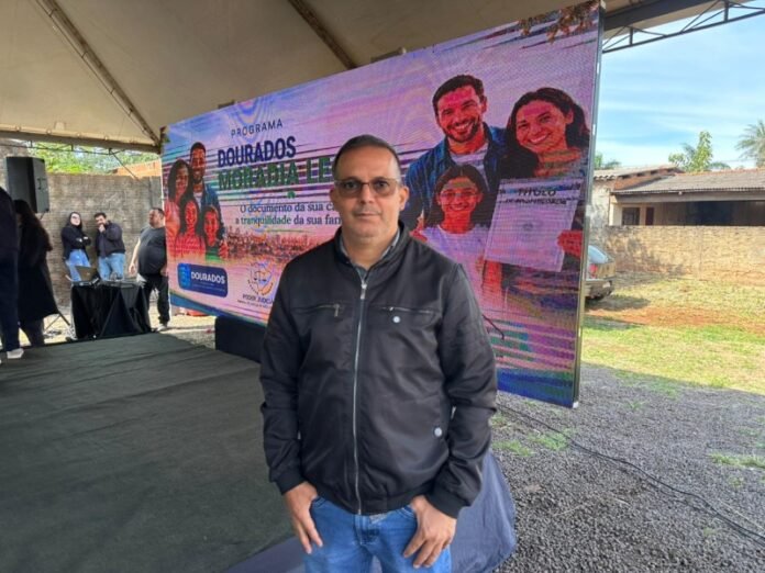 Man standing in front of a colorful event banner under a tent, promoting a community program in Dourados.