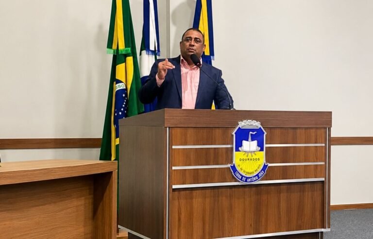 Man in a pink shirt and navy blazer speaks from a wooden podium with a blue-yellow city crest, flags behind him.