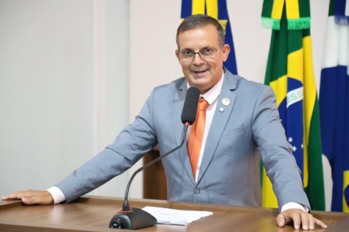 Man in a gray suit and orange tie speaks at a wooden podium with a microphone, national flags in the background.