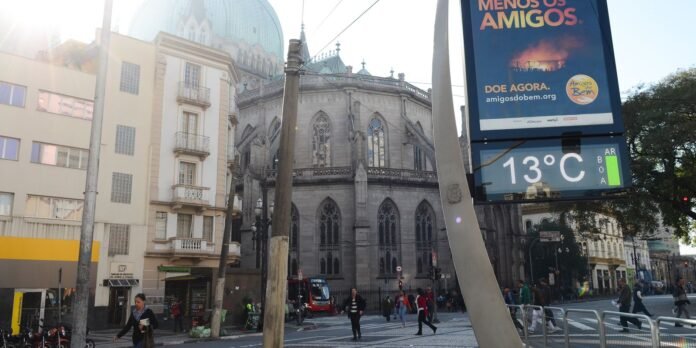 A city street with a Gothic-style cathedral, pedestrians crossing, and a tall digital sign showing 13°C and an ad banner nearby.