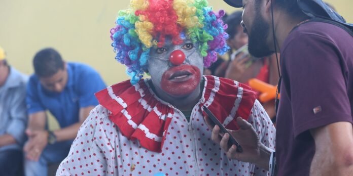 Clown with a rainbow curly wig, gray face paint, red nose and red polka-dot outfit talks to a man holding a smartphone nearby.