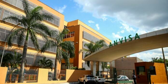 Exterior view of the Anvisa building with an orange facade, palm trees, and a curved walkway canopy at the entrance.