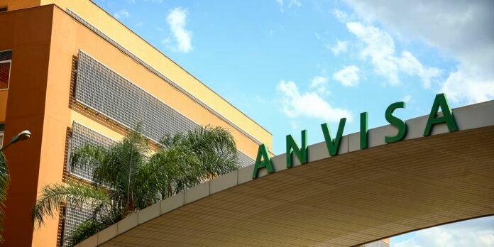 Orange building with a green ANVISA sign over a beige canopy and palm trees against a blue sky.