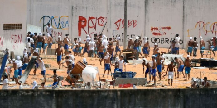 Crowded shoreline with many shirtless men in blue shorts and white head coverings, building shelters amid debris near a graffiti-covered wall.