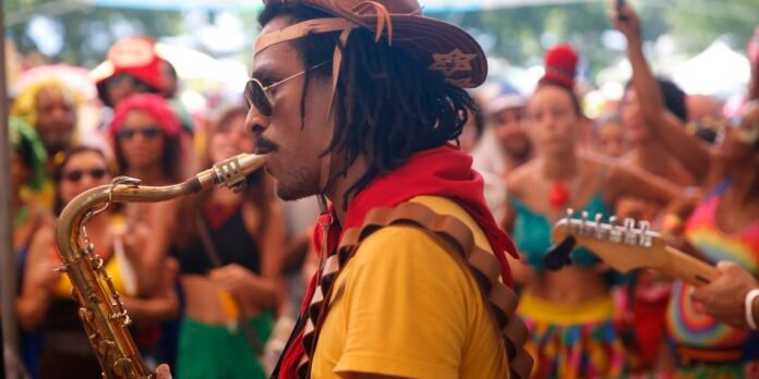 Saxophonist in a yellow shirt and brown hat plays a brass sax at a crowded outdoor festival.