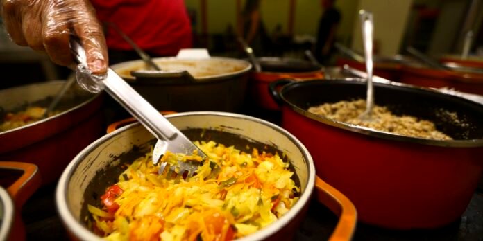Person wearing a glove uses a large spatula to stir chopped sautéed vegetables (cabbage, peppers) in a pot at a busy kitchen/buffet line.