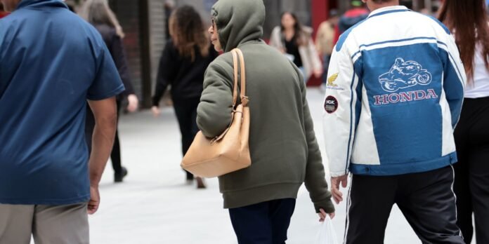 Back view of a person in an olive-green hoodie carrying a tan handbag while walking in a busy street, with a Honda-clad person nearby.