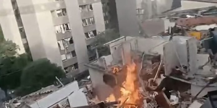 Fire and debris erupting from a collapsed building in an urban area, with apartment blocks nearby