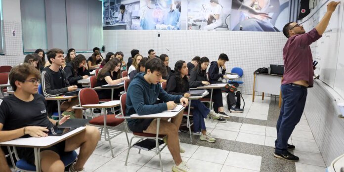 A teacher writes on a whiteboard at the front of a classroom while students sit at desks and take notes.