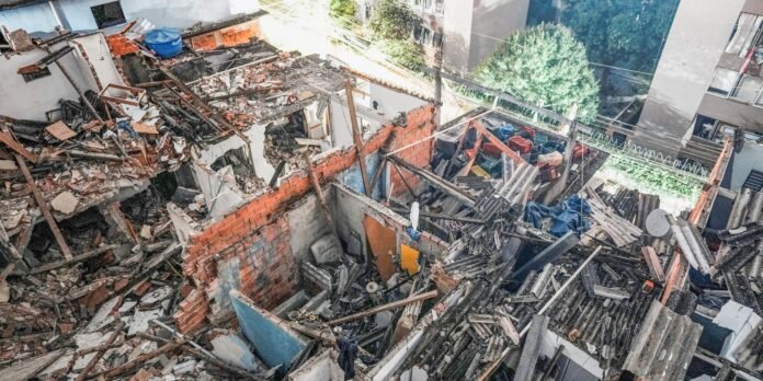 Wide view of a collapsed building with red brick walls, tangled metal beams, and concrete rubble in an urban courtyard.