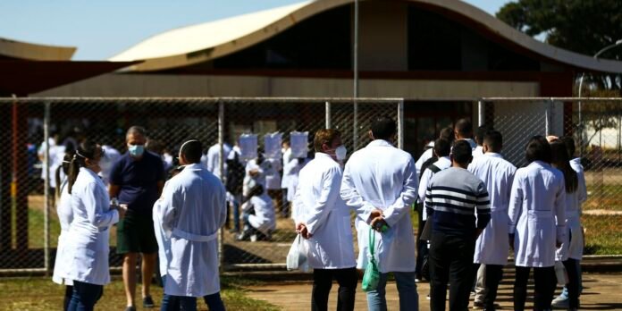 Group of people in white lab coats and masks standing outside a fenced area near a modern building.